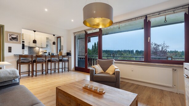 A modern living room with a wooden coffee table, a sofa, a chair, and a view of a kitchen and outside landscape through large windows.