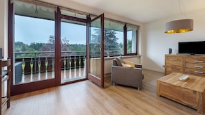 A spacious living room with a large sliding glass door leading to a balcony, a grey sofa, a wooden coffee table, and a TV on a wooden cabinet.