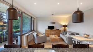 A modern living room with a sofa, coffee table, and dining table. There is a television, a lamp, and a wooden cabinet.