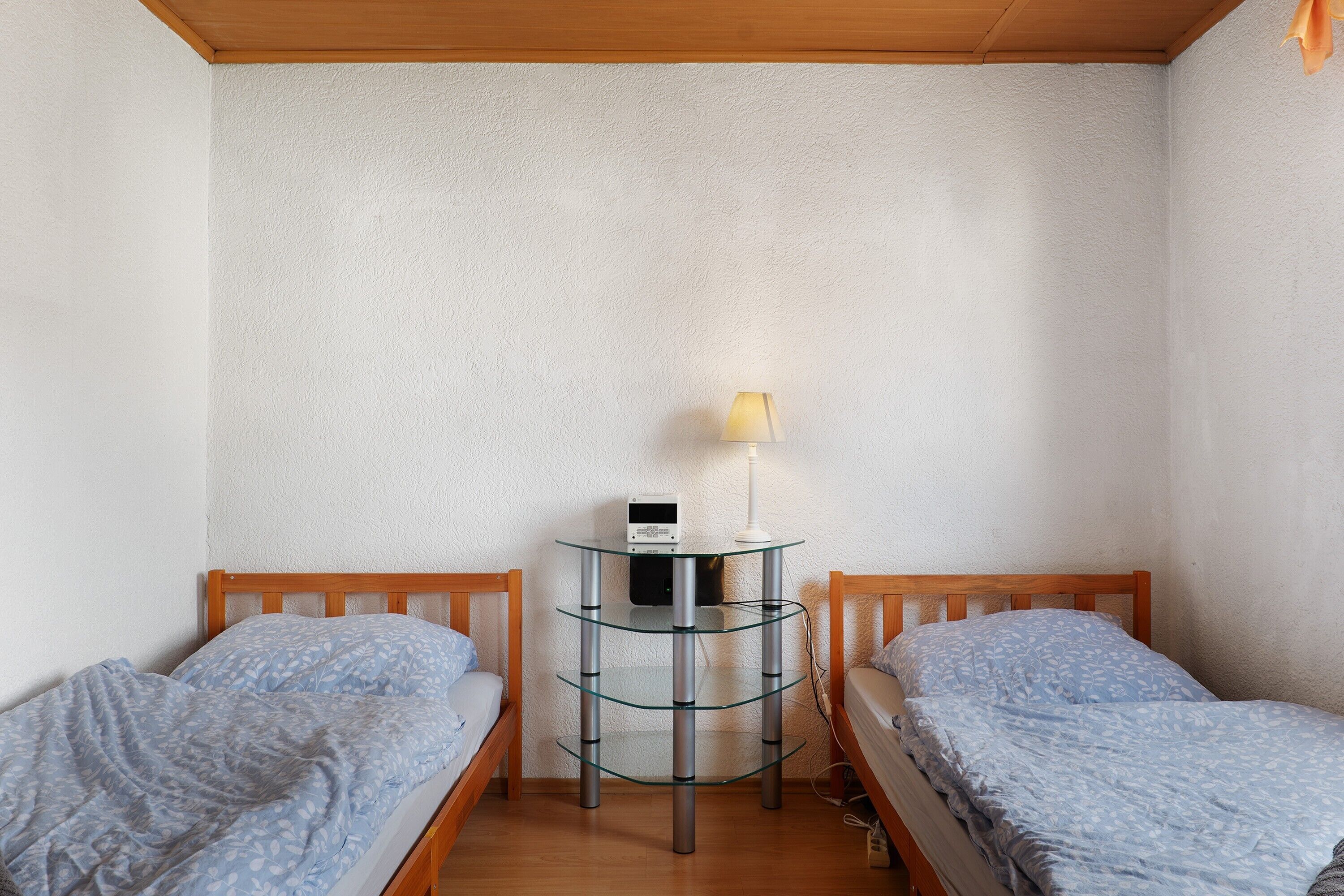 A small room with two wooden beds, a glass table with a lamp, and a white wall.