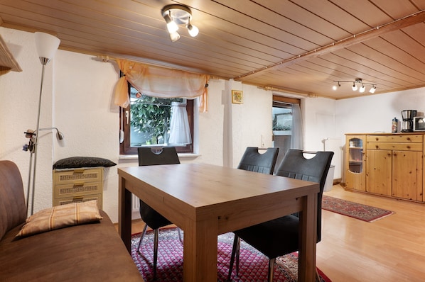 A dining area with a wooden table and chairs, a sofa, a lamp, a window with curtains, and a kitchen area with cabinets and appliances.
