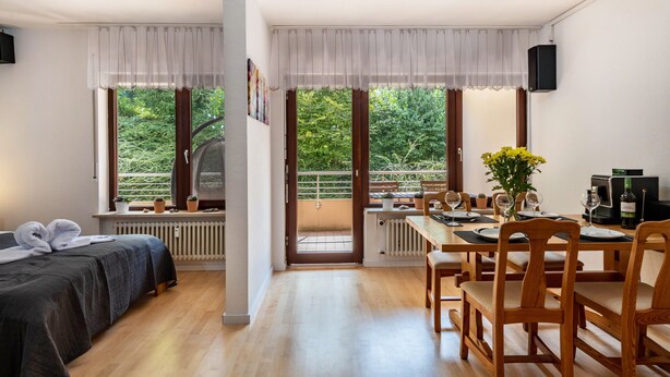 A room with a bed, a dining table with chairs, a radiator, a window with curtains, and a balcony with greenery. - Apartment 'Zwergenbau' with Shared Garden, Balcony, and Wi-Fi (Badenweiler)