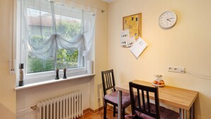 A small room with a window, a wall clock, a radiator, a table with chairs, and a bulletin board with papers.