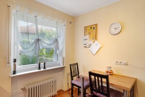 Una habitación pequeña con ventana, un reloj de pared, un radiador, una mesa con sillas y un tablón de anuncios con papeles.