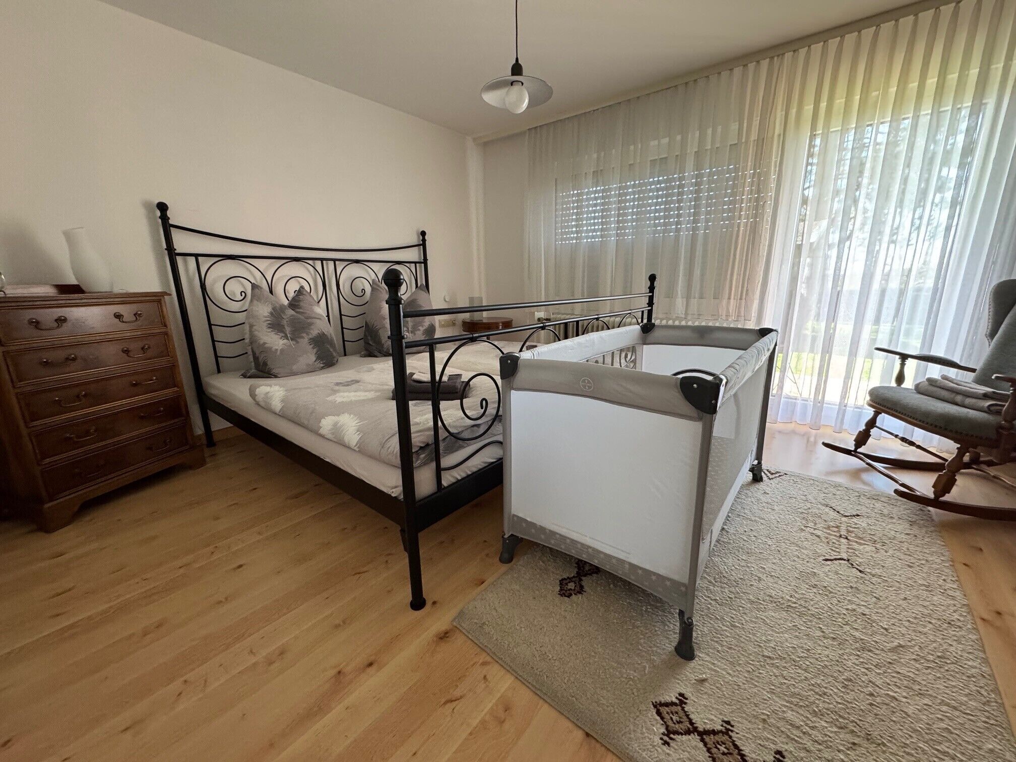Ein Schlafzimmer mit einem Bett, einer Kommode, einem Schaukelstuhl und einem Fenster mit Vorhängen.