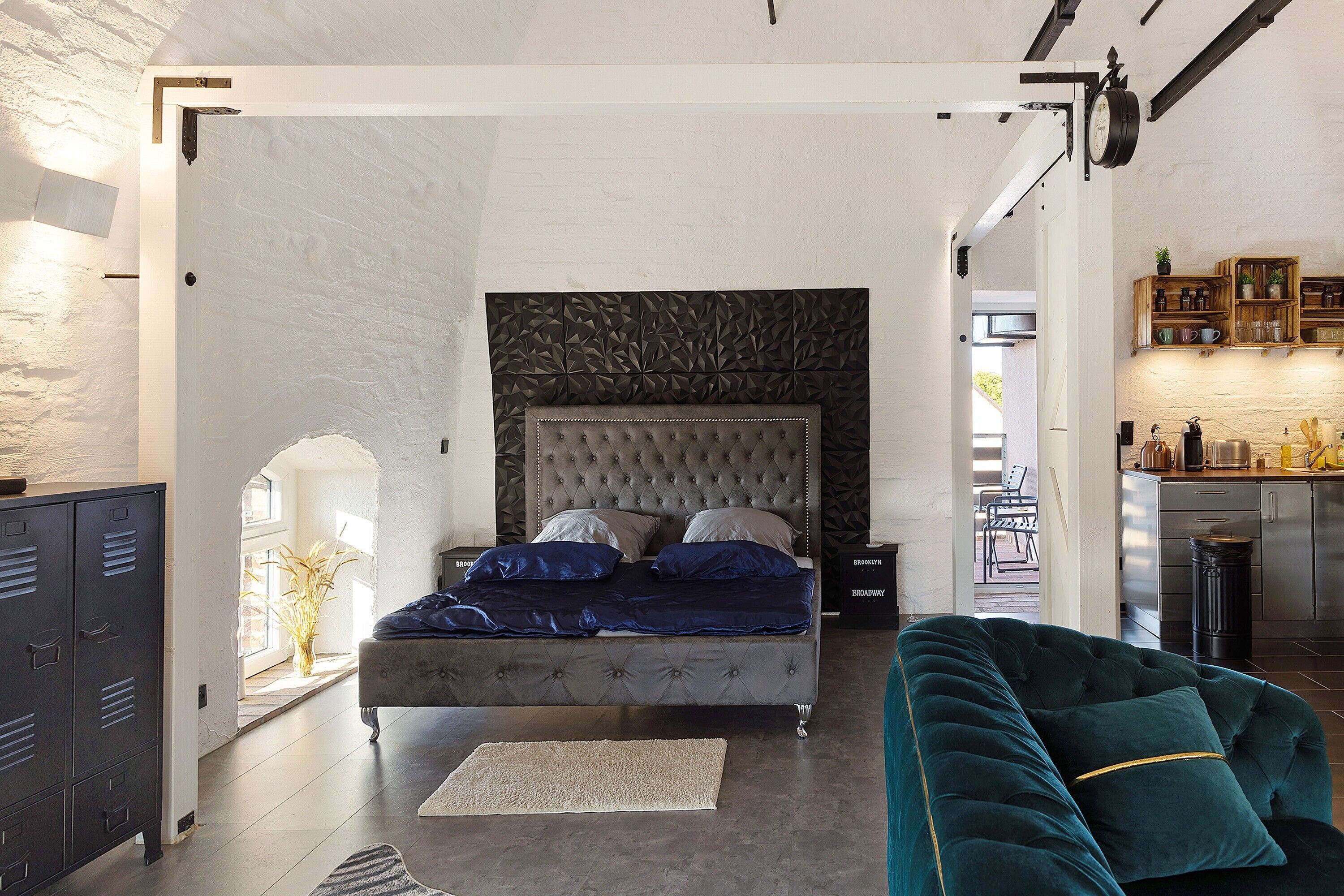 A bedroom with a tufted headboard, a bed with blue bedding, a green sofa, a wooden cabinet, and a kitchen area visible through an open doorway.