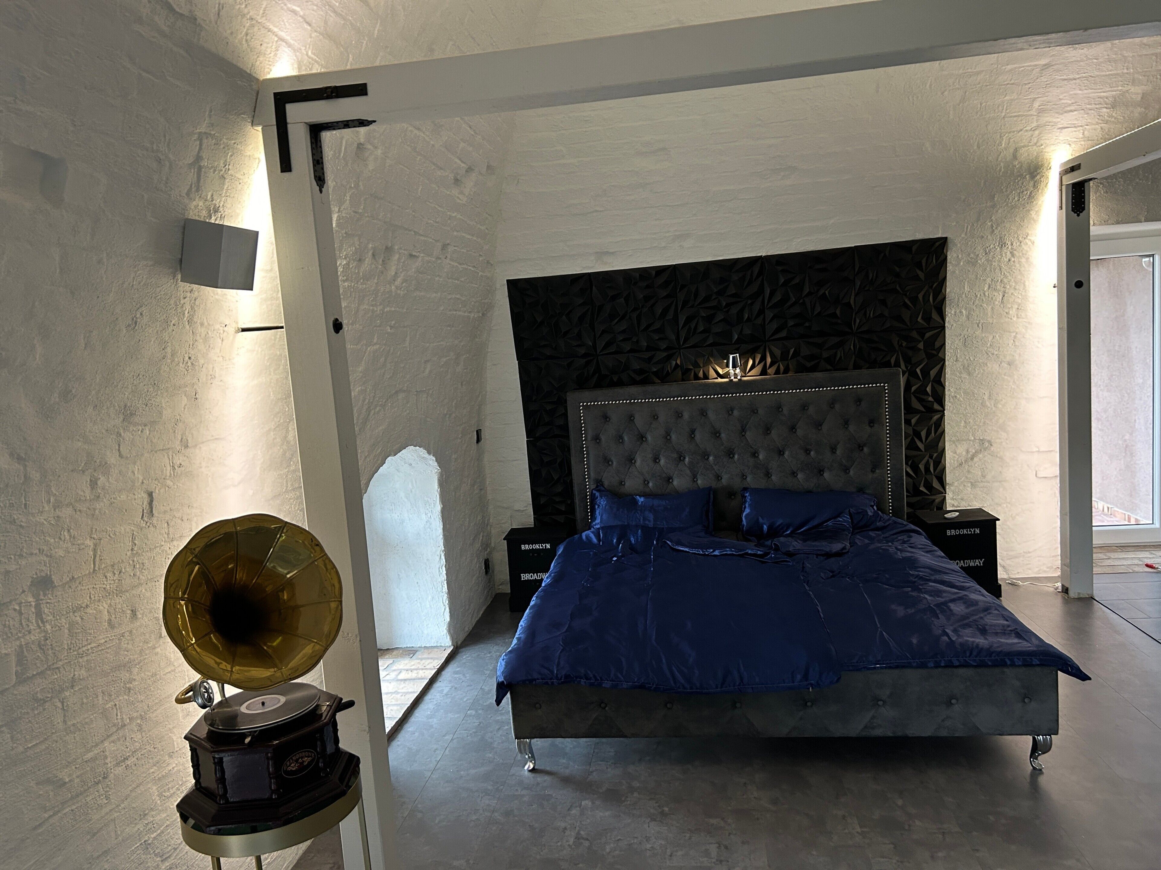 Une chambre à coucher avec une tête de lit capitonnée, un lit avec une literie bleue, un gramophone posé sur un support et un mur foncé à la texture marquée.