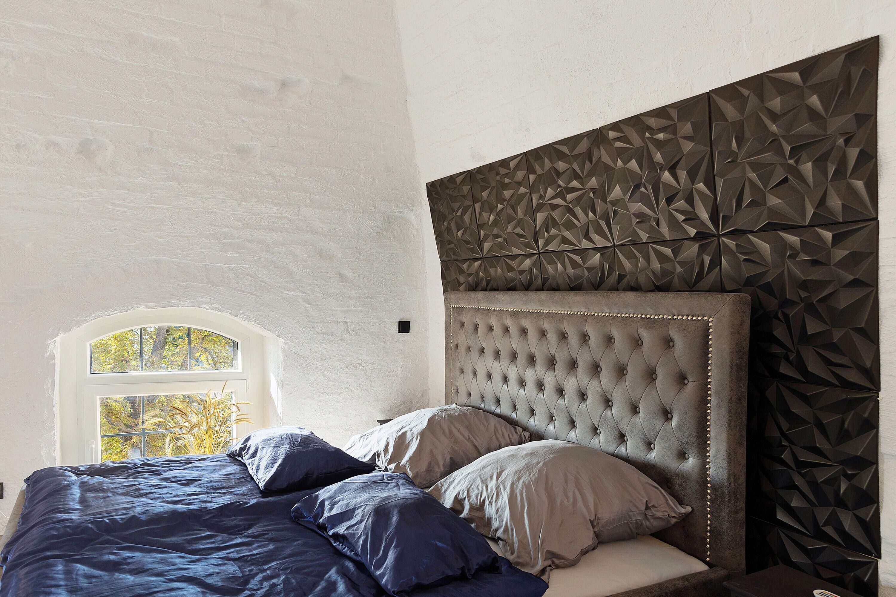 Une chambre à coucher avec une tête de lit capitonnée, une fenêtre donnant sur des arbres et un mur orné d’un motif géométrique.
