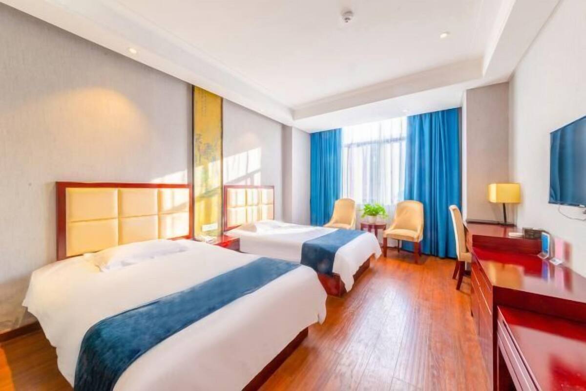 A hotel room with two beds, a desk, a chair, a TV, and a window with curtains.