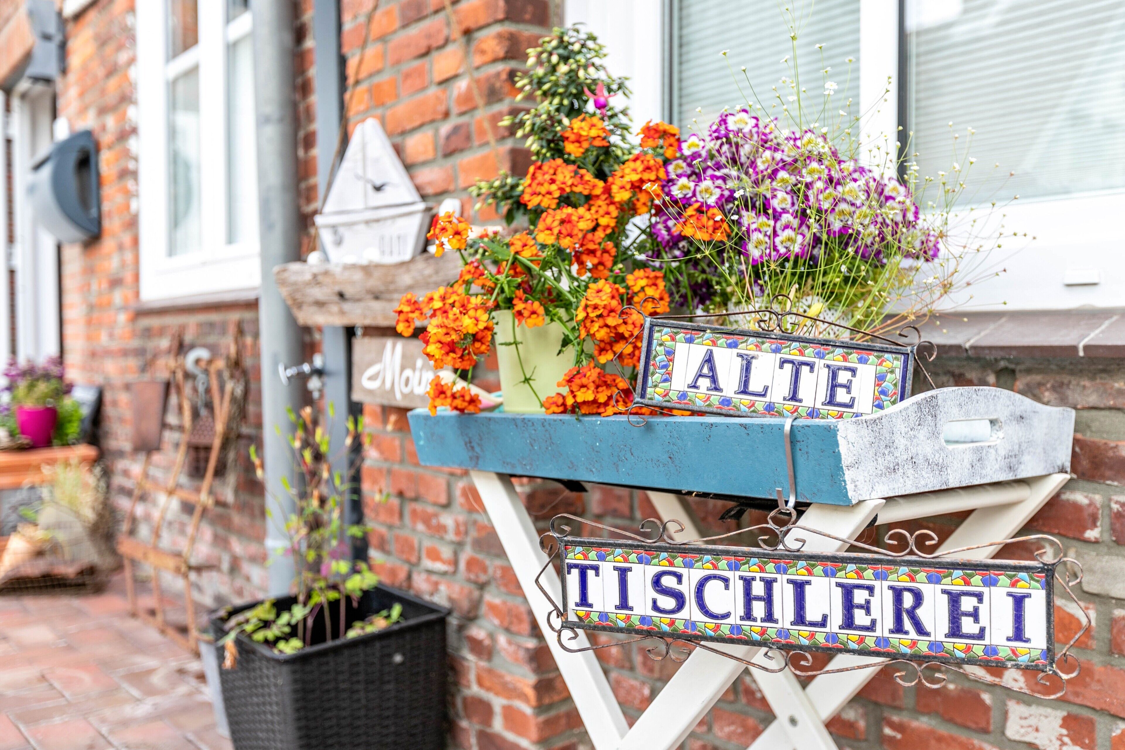En blå skylt med orden ”ALTE” och ”TISCHLEREI” på, fäst på ett vitt stativ med en blomsterdekoration.