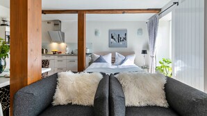 A modern bedroom with a sofa, a bed with white bedding, a kitchen in the background, and a large window with curtains.