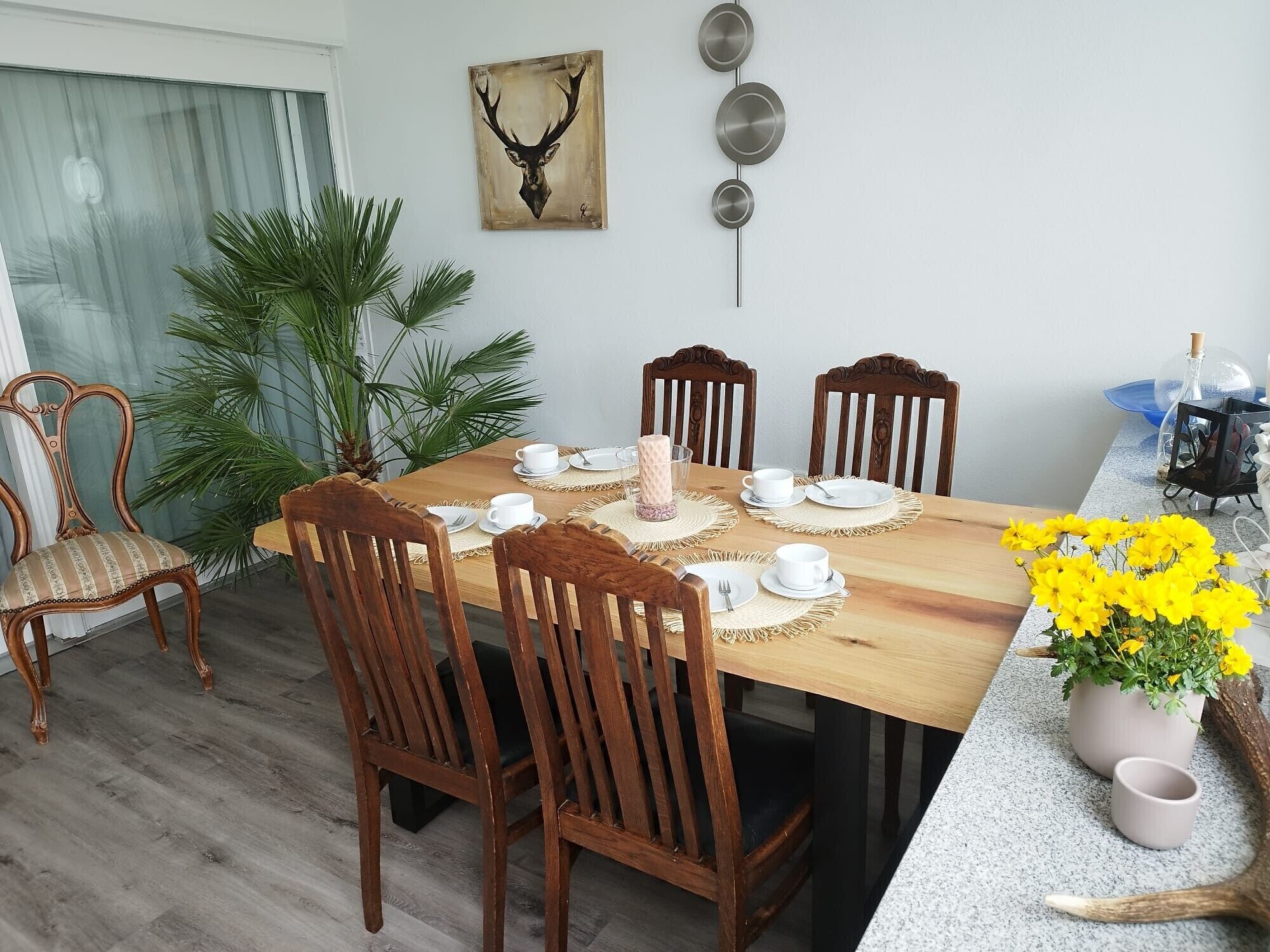 A dining area with a wooden table, four chairs, a vase of yellow flowers, and a wall-mounted deer head painting.