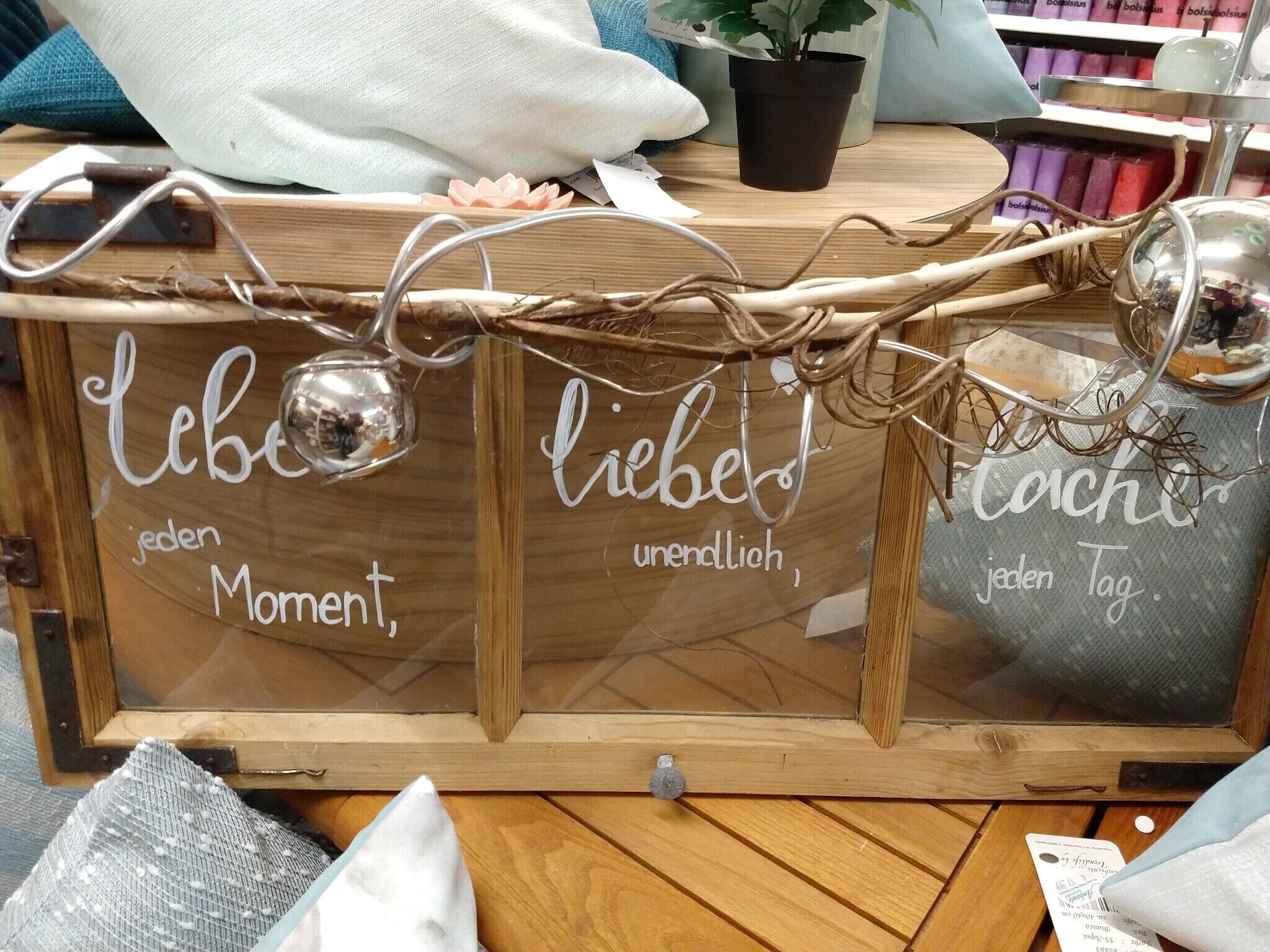 Una vitrina de madera con la inscripción 'Liebe jeden Moment, liebe jeden Tag' y elementos decorativos como ramas y una planta pequeña.