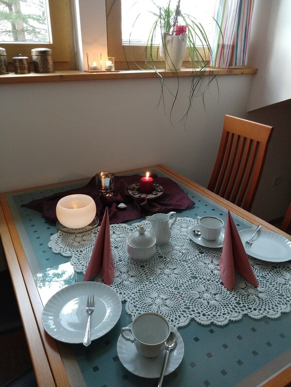 A dining table set with plates, cups, and a candle, with a window view and a plant on the sill.
