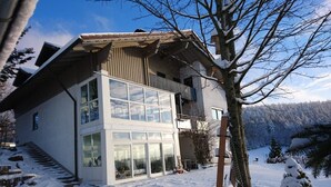 A modern two-story building with large windows and a balcony, surrounded by snow-covered trees and a mountainous backdrop. - Grabmeier holiday flat - number 3’ with mountain view, balcony and Wi-Fi (Grainet)