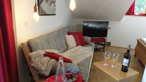 A cozy living room with a sofa, a flat-screen TV, a wooden table with wine glasses, and a red curtain. - Grabmeier holiday flat - number 3’ with mountain view, balcony and Wi-Fi (Grainet)