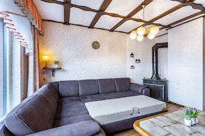 Un salon moderne avec un canapé d’angle, une petite table, une horloge murale, une cheminée et un plafond avec des poutres apparentes.
