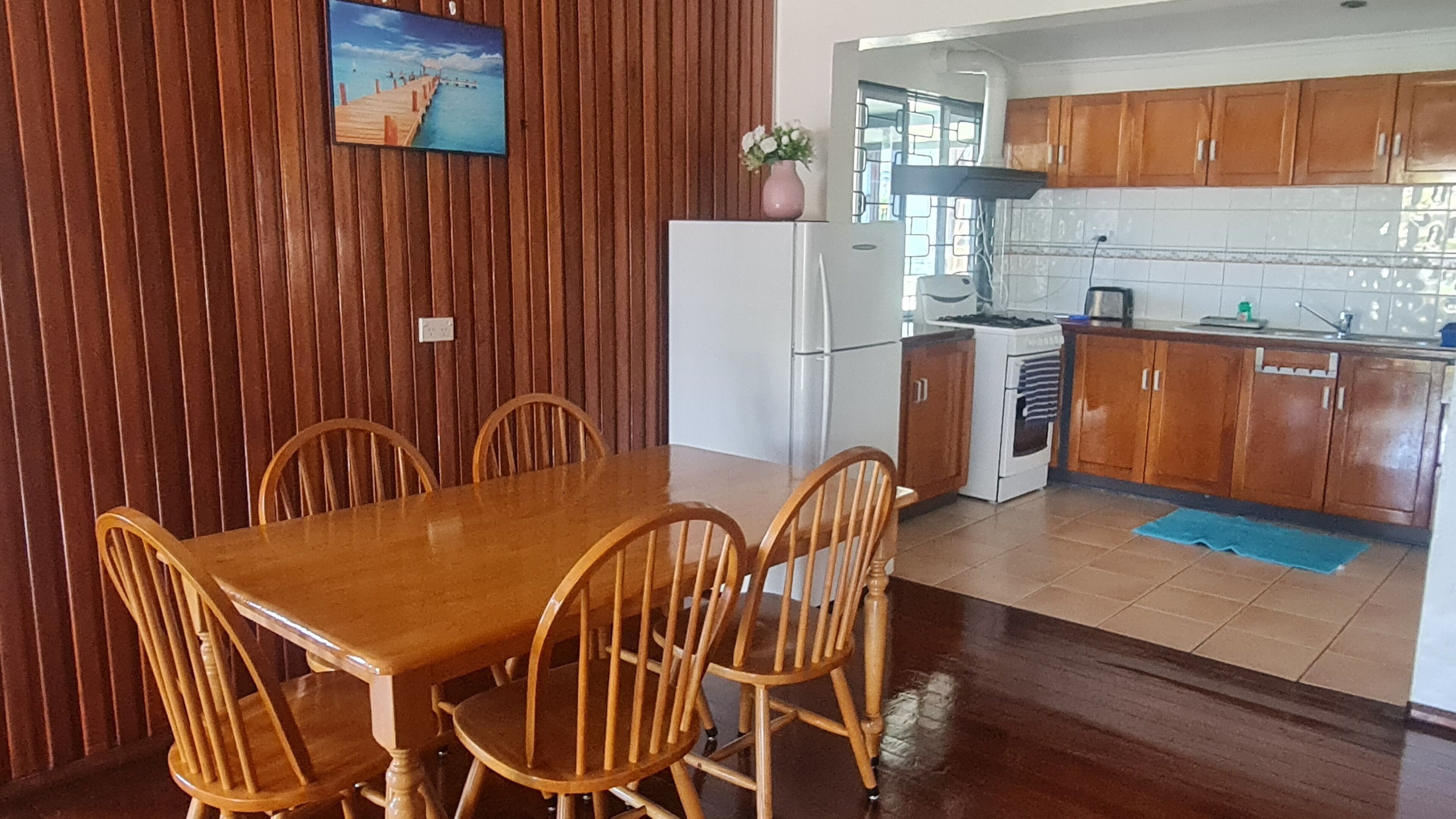 Een eethoek met een houten tafel en stoelen, een koelkast, een fornuis, en een keuken met houten kastjes en een betegelde spatwand.