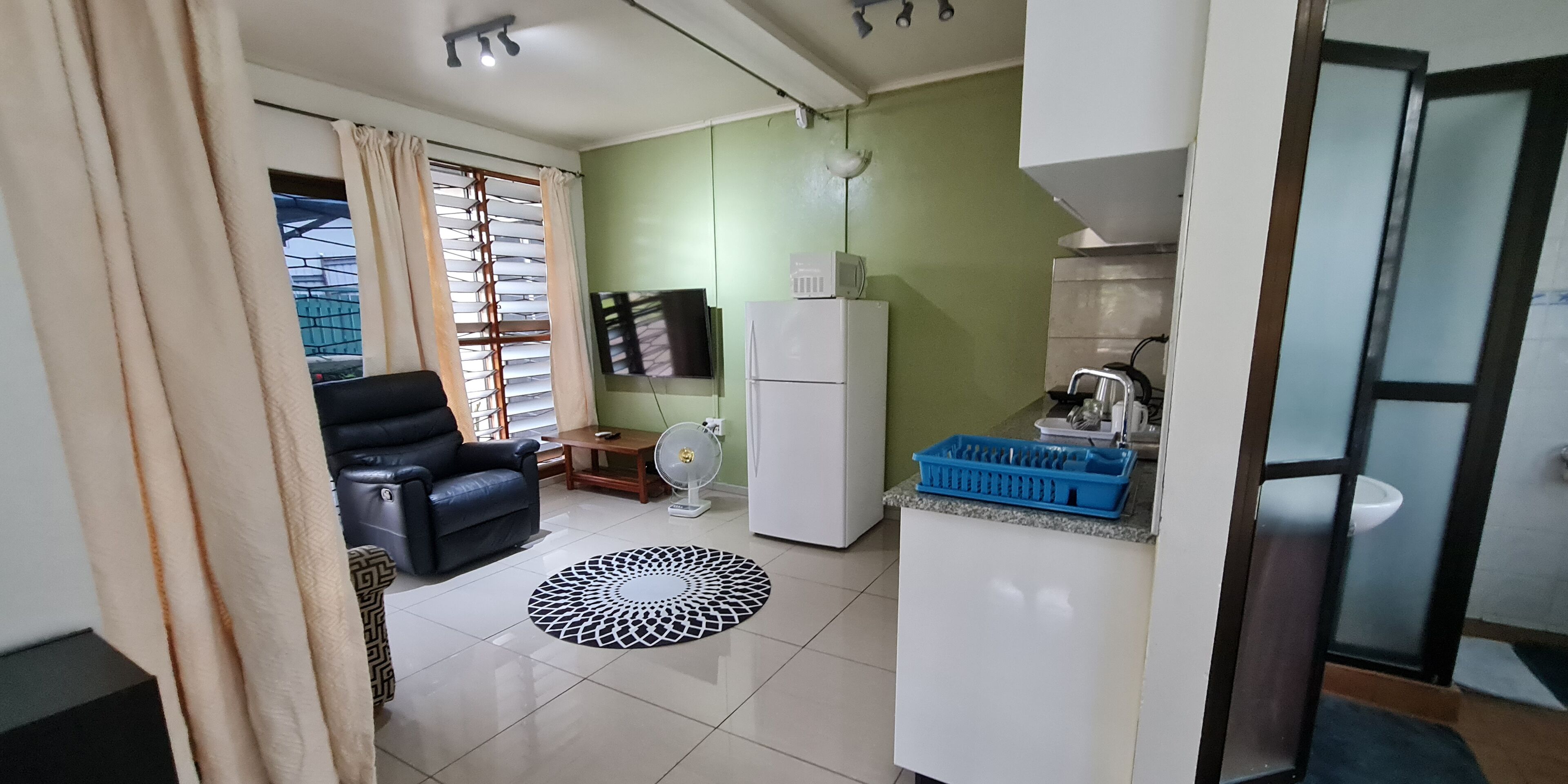 Una sala de estar con un sofá de cuero negro, una mesa de centro de madera y una alfombra estampada. Hay un televisor montado en la pared y un refrigerador blanco.