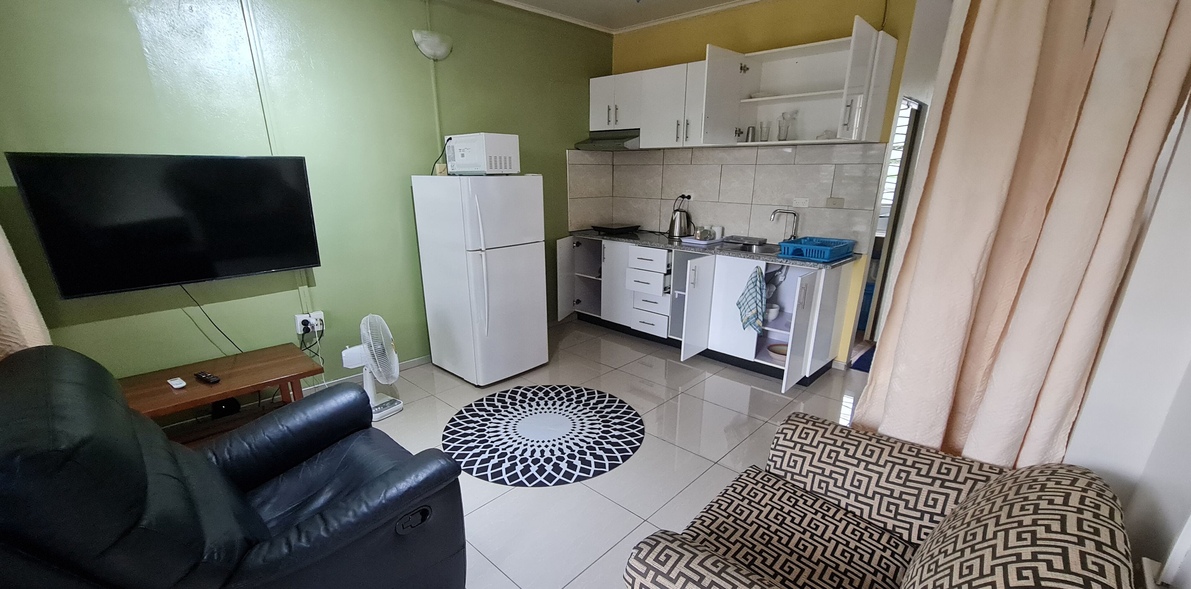 A compact living space with a flat-screen TV, a refrigerator, a kitchenette, a sofa, and a patterned rug.