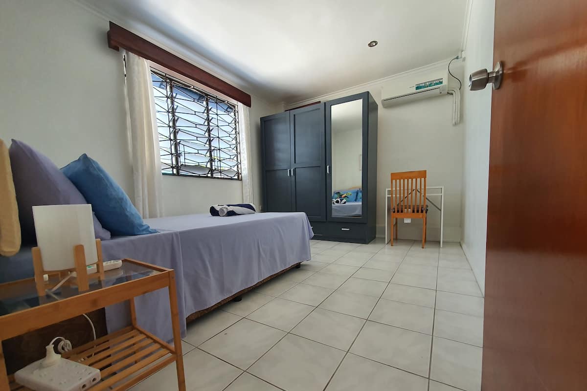 A bedroom with a bed, a wooden nightstand, a chair, a wardrobe, and a window with a metal grill.