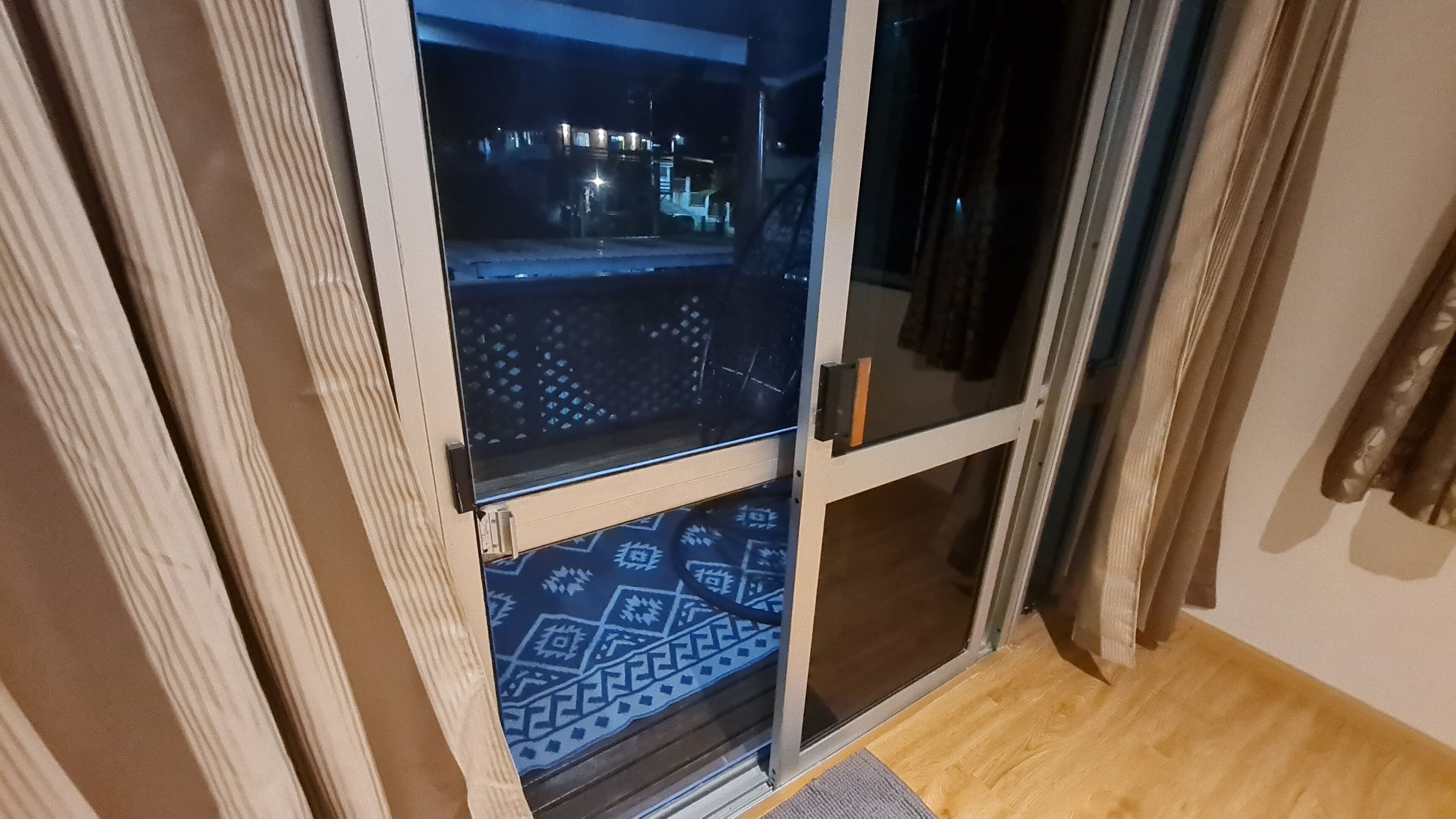 A room with a sliding glass door leading to a balcony. The door has a metal handle and a glass panel with a geometric pattern. The balcony has a wooden floor and a patterned mat.