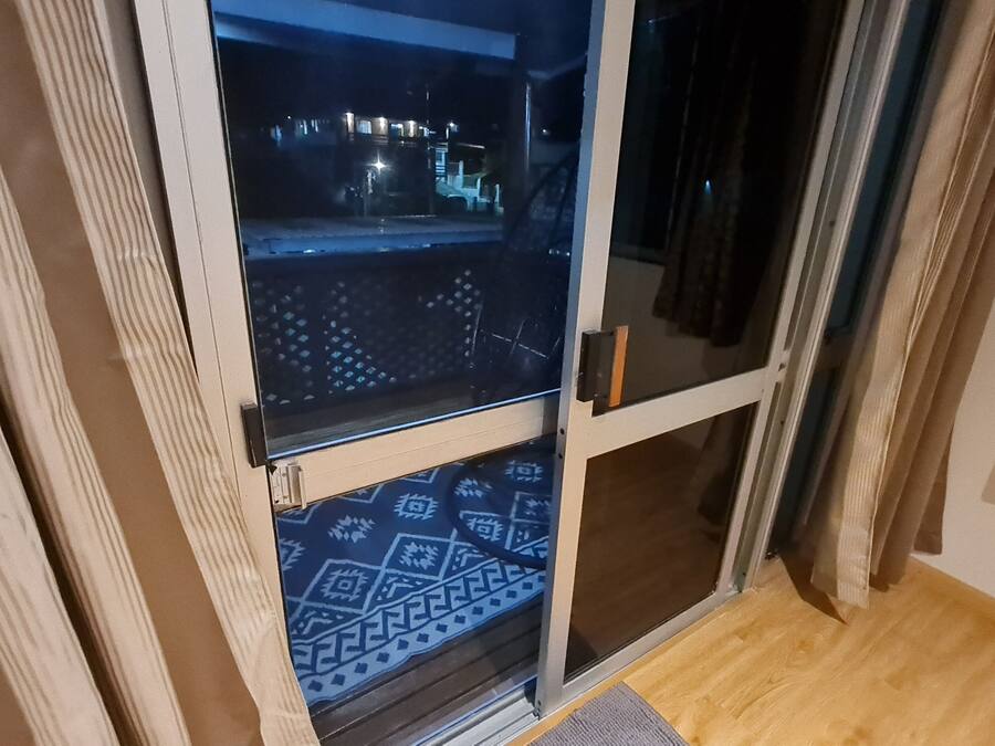 A room with a sliding glass door leading to a balcony. The door has a metal handle and a glass panel with a geometric pattern. The balcony has a wooden floor and a patterned mat.