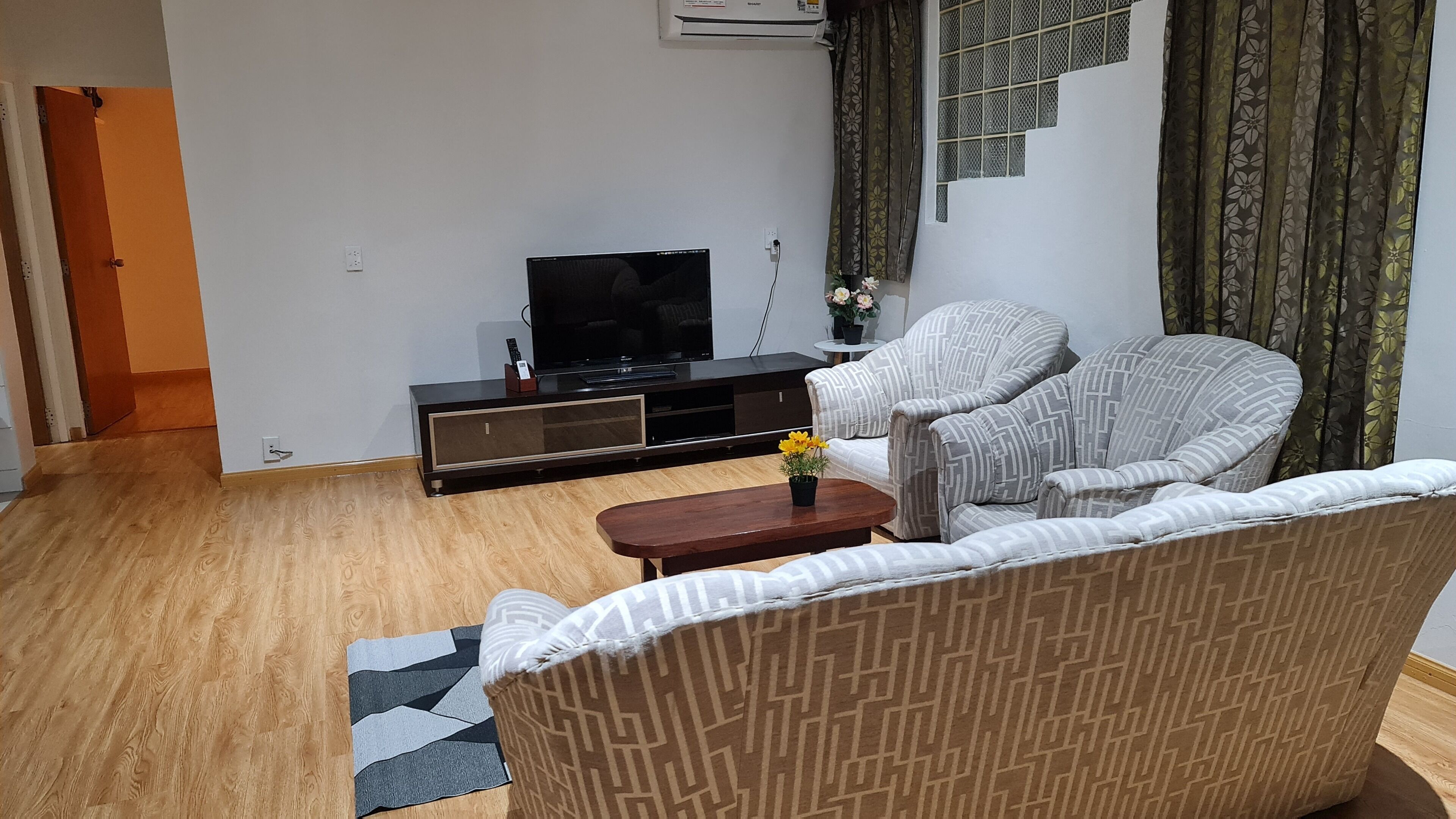 A living room with a sofa, a coffee table with a flower vase, a TV on a stand, and a staircase with patterned curtains.