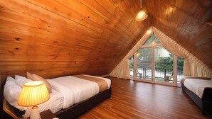 A cozy attic bedroom with wooden ceilings, a large window with curtains, and a bed with a bedside lamp.