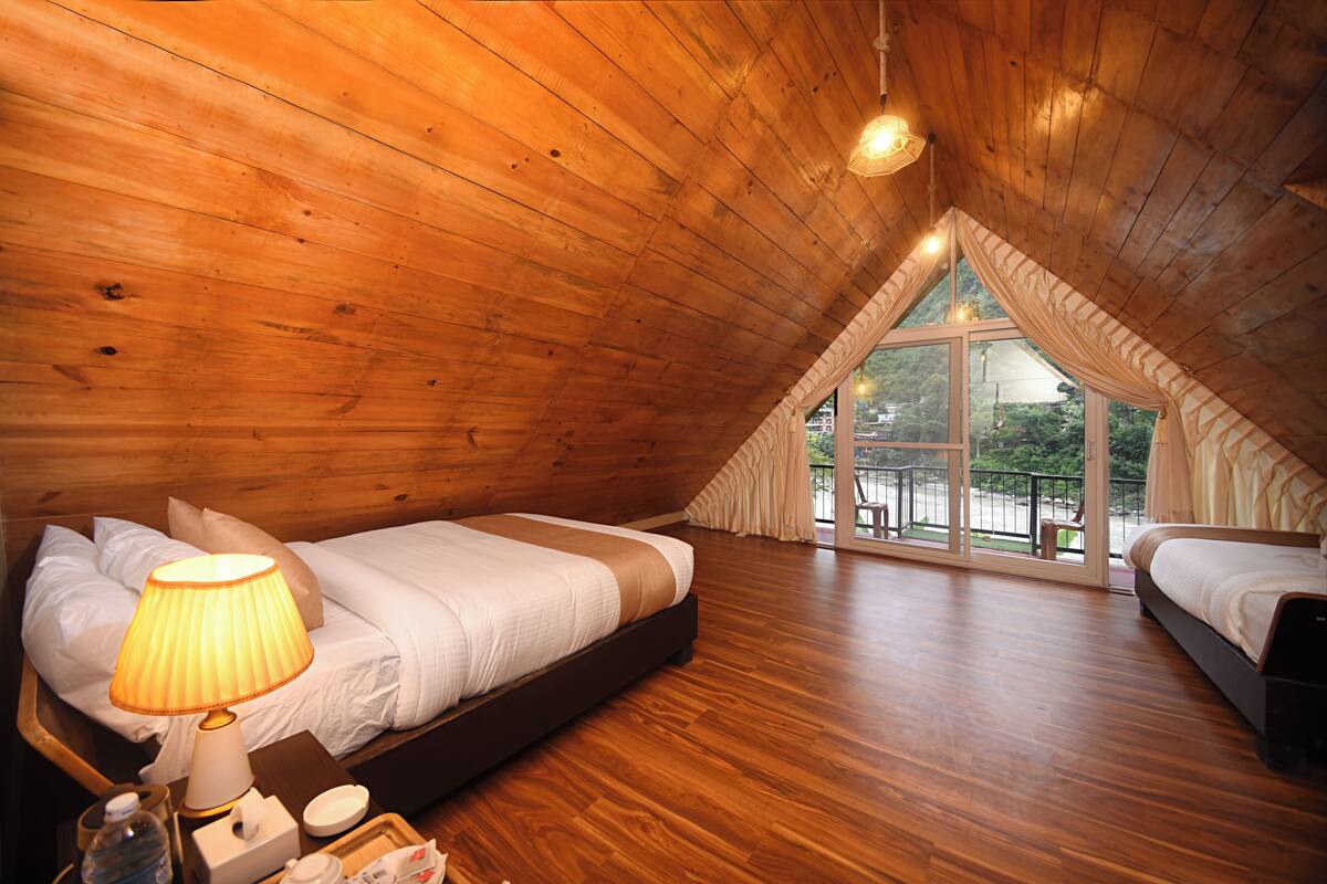 Un dormitorio acogedor en el ático, con techos de madera, un ventanal con cortinas y una cama con lámpara auxiliar.