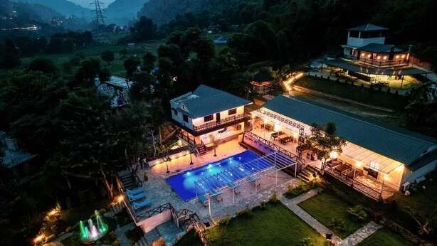 A two-story building with a swimming pool, surrounded by greenery and a hilly landscape.