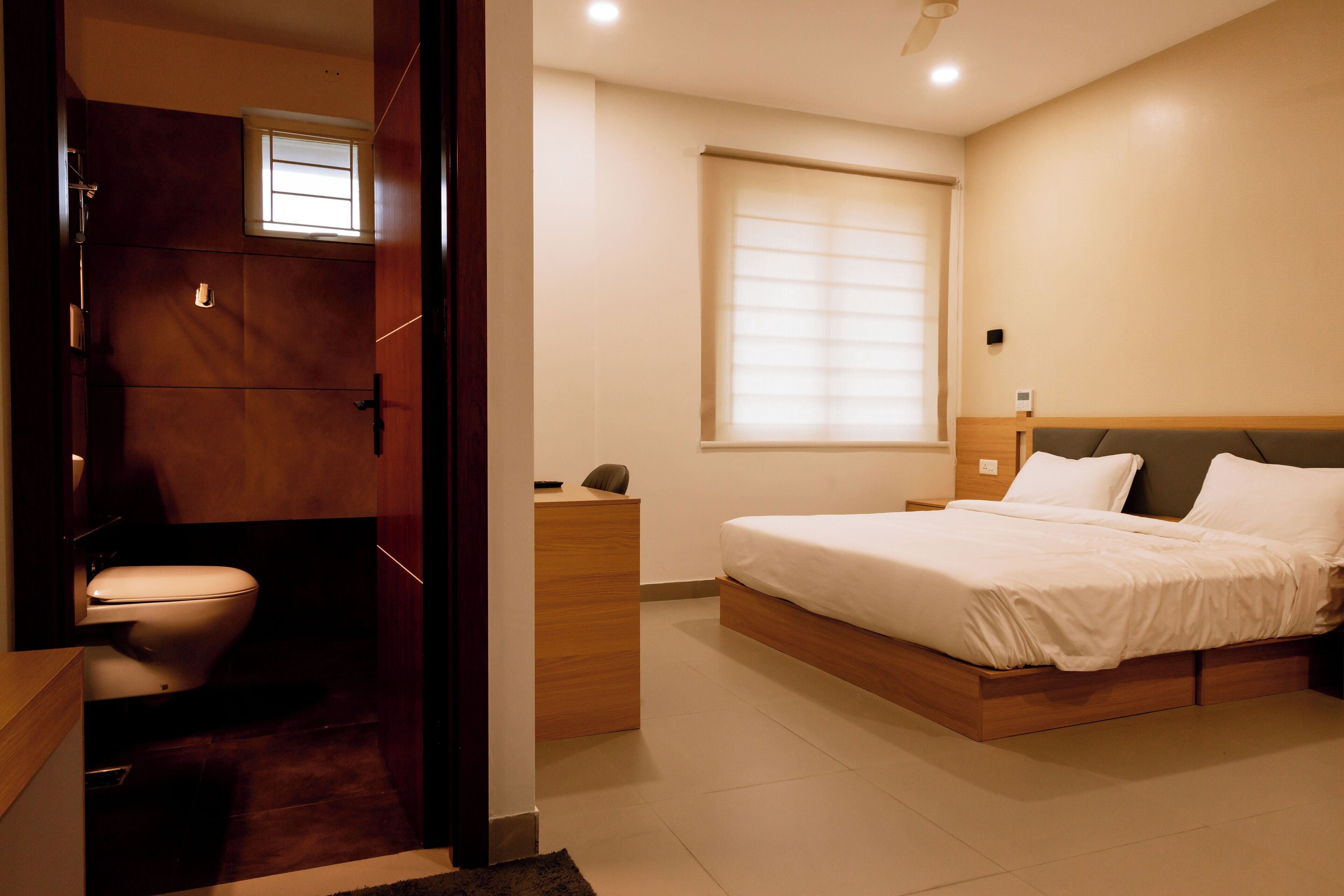 A bedroom with a wooden bed, a bedside table, a nightstand, a window with blinds, and a door leading to a bathroom.