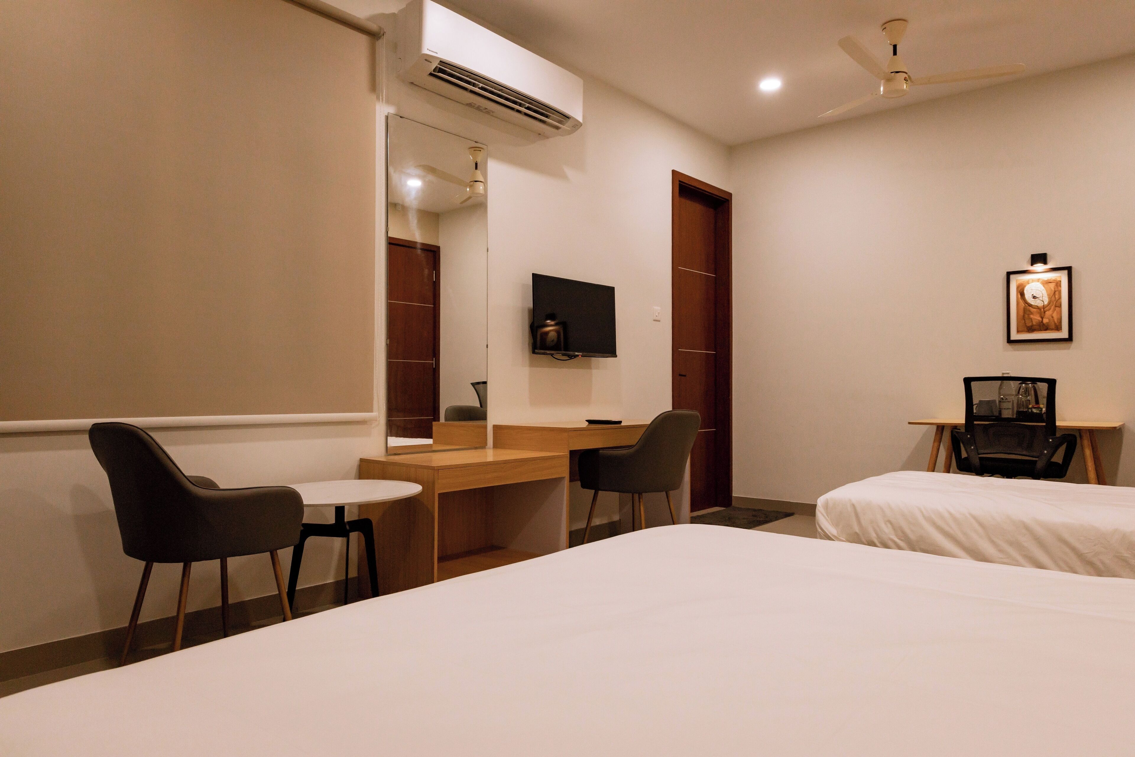 A hotel room with a bed, a desk with two chairs, a TV, and an air conditioning unit.