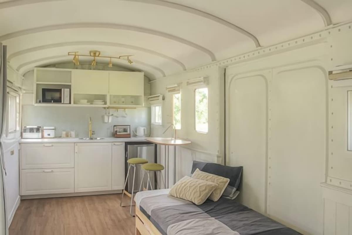 A compact kitchen with a microwave, sink, and stools, a bed with pillows, and a window providing natural light.