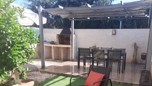 A patio with artificial grass, two black lounge chairs with red cushions, a glass coffee table, and a barbecue grill.