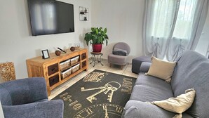 A living room with a grey sofa, a wooden side table with baskets, a potted plant, a small armchair, and a flat-screen TV mounted on the wall.