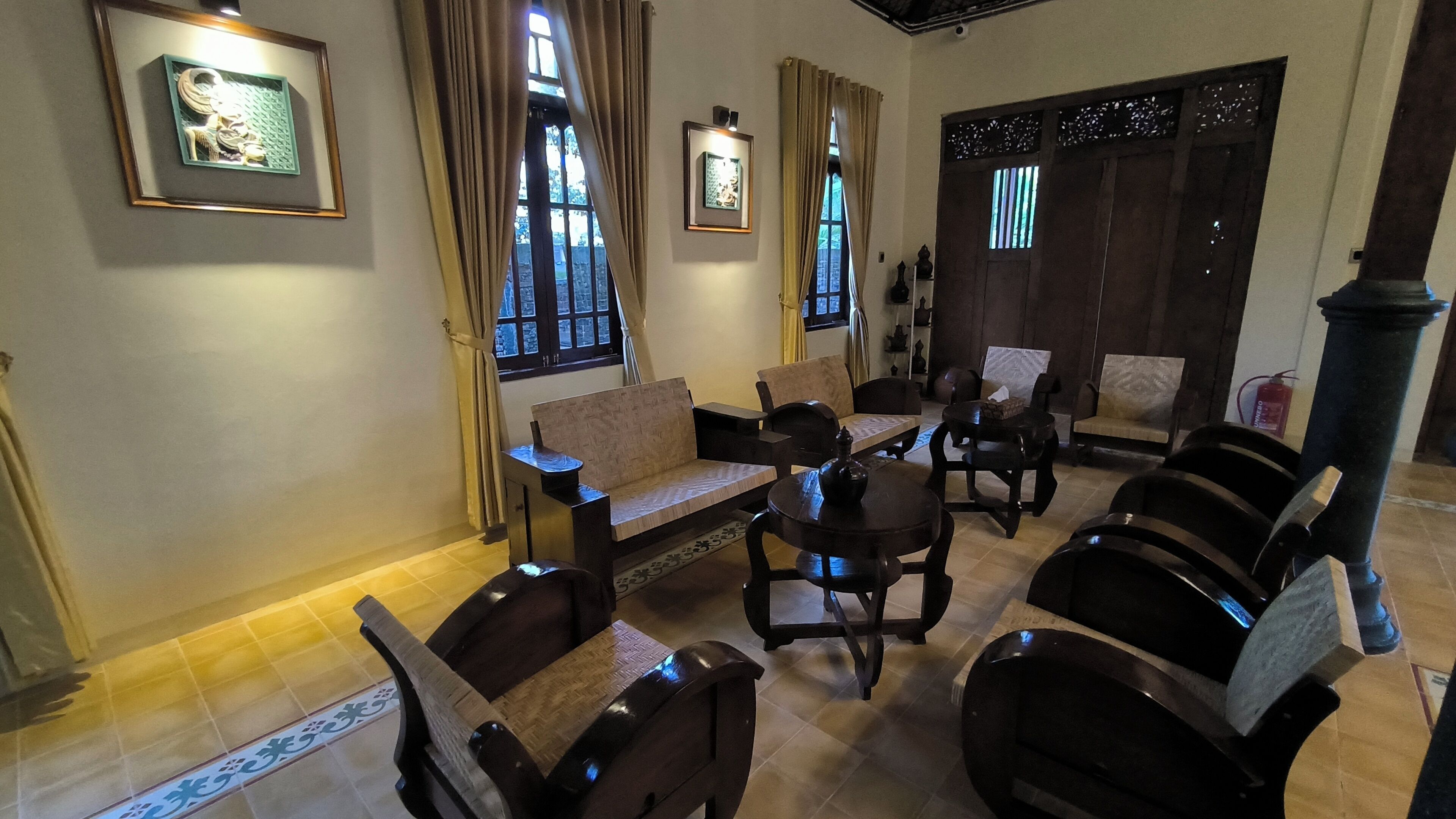 Habitación de estilo tradicional con muebles de madera, incluyendo sofás y sillones, organizados alrededor de una mesa central. Se observan obras de arte enmarcadas en las paredes, y la habitación cuenta con ventanas grandes con cortinas.