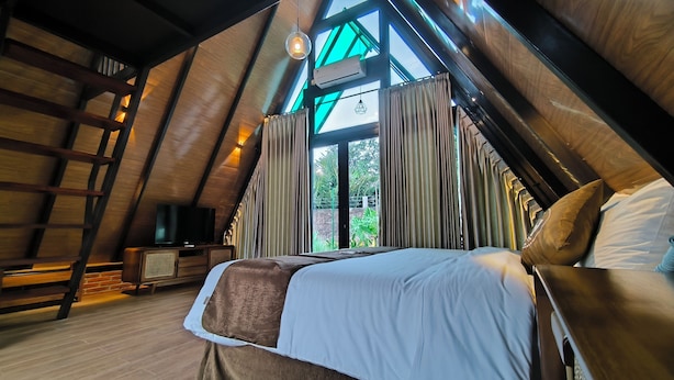 Une chambre avec un plafond en pente, un lit avec des oreillers, un téléviseur posé sur un meuble en bois, et une vue sur la verdure à l’extérieur.