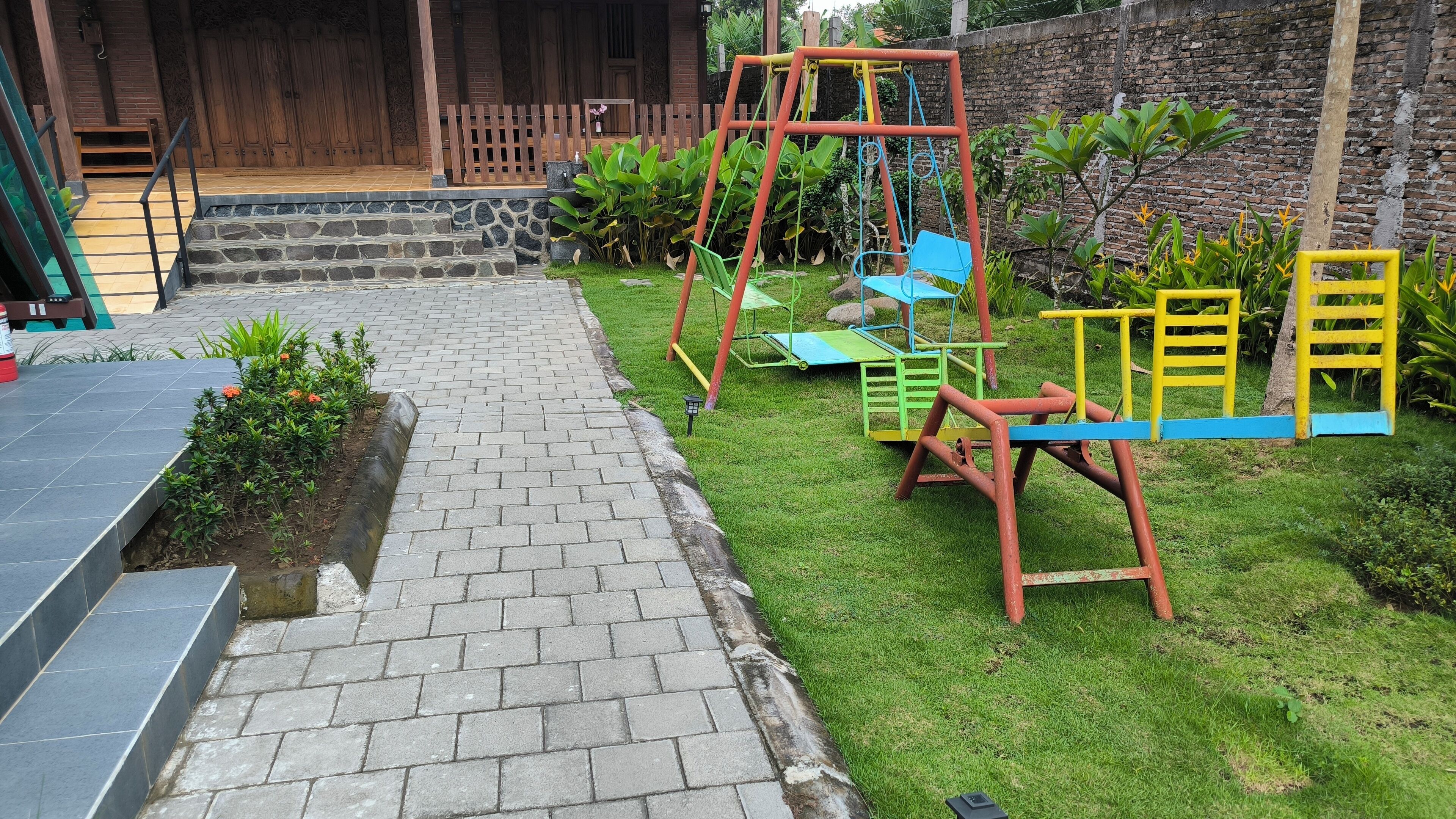 Un chemin pavé mène à une porte en bois, à une balançoire et à un petit jardin agrémenté de plantes et d’un banc.