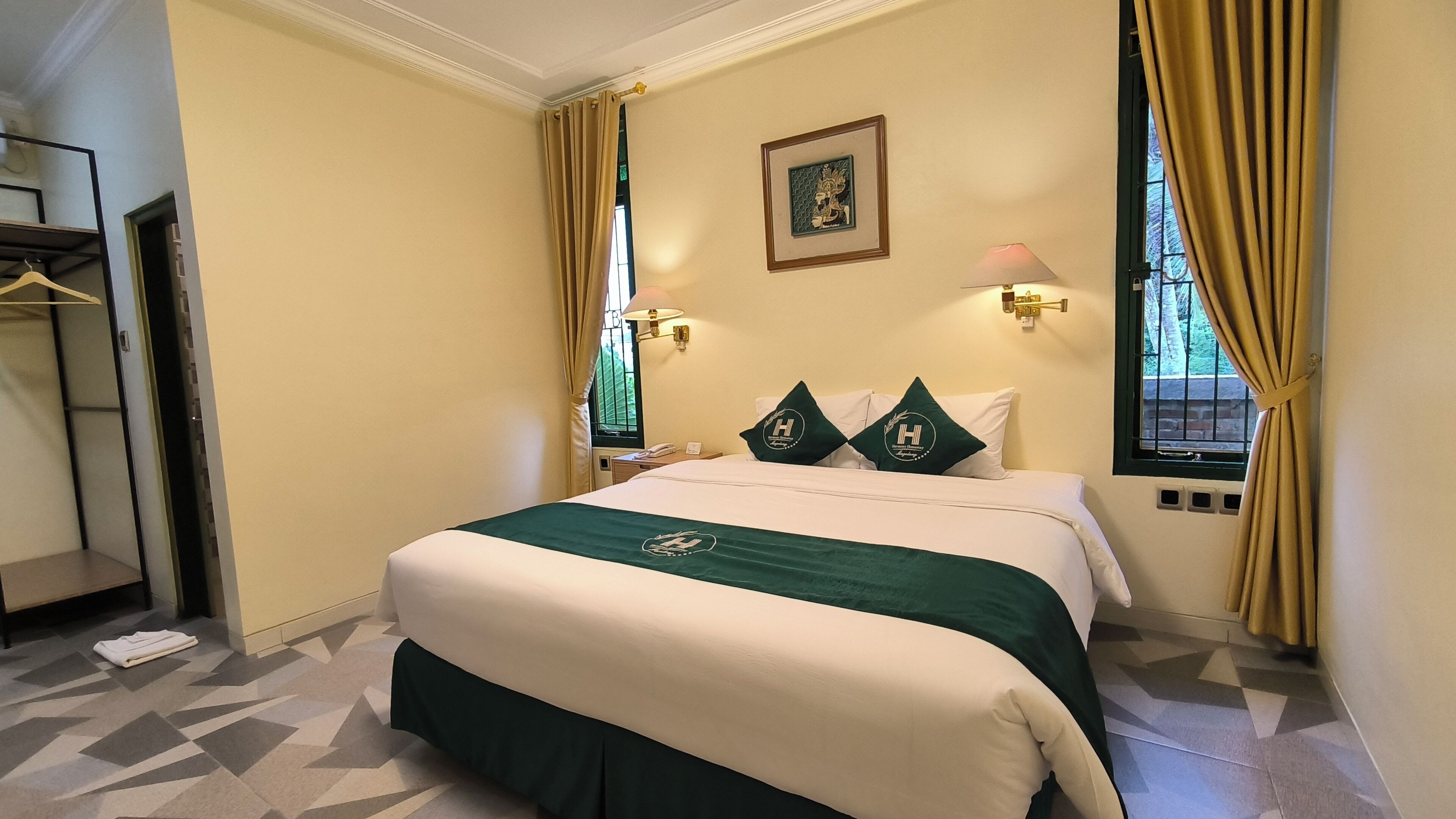 Una habitación de hotel con una cama bien hecha, una mesita de noche con lámpara, un cuadro en la pared y una ventana con cortinas.