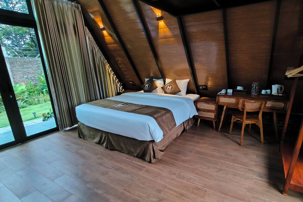 Ein Schlafzimmer mit Bett, Schreibtisch und Stuhl. Ein Fenster mit Vorhängen bietet Blick auf grüne Vegetation.