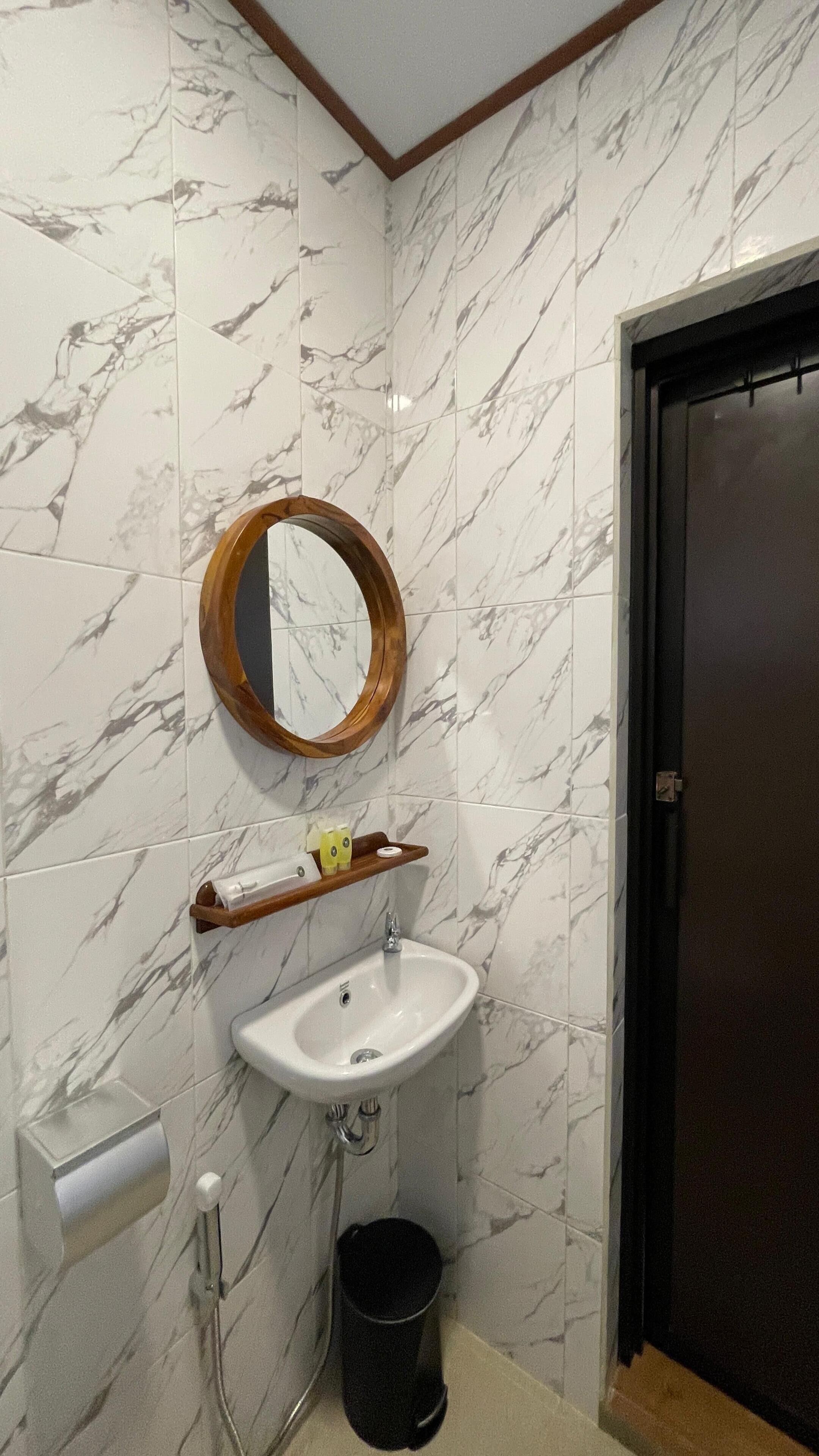 Ein Badezimmer mit Marmor-Spritzschutz, einem weißen Waschbecken, einem Holzbodenregal, einem Spiegel, einer Tür und einem Mülleimer.