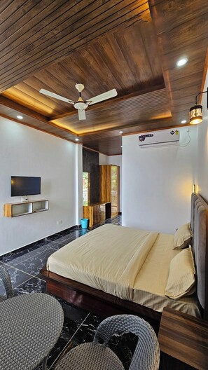 Un dormitorio moderno con techo de madera, un televisor de pantalla plana, una cama con ropa de cama color beige y un rincón de descanso con sillas de mimbre.