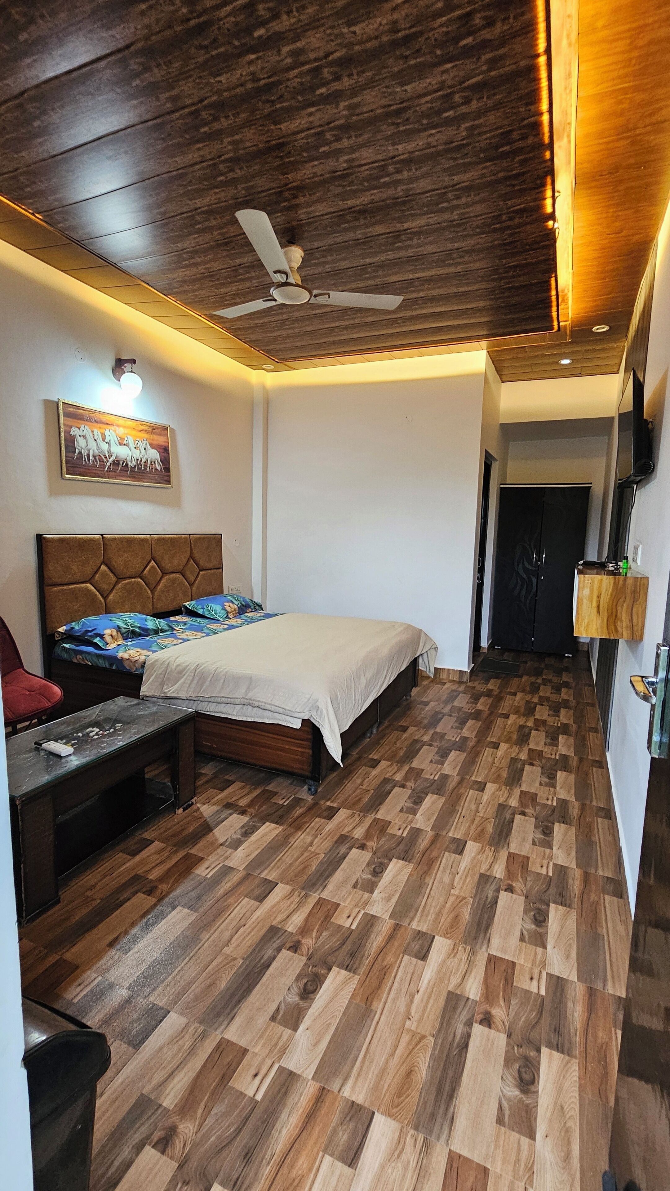 A bedroom with a wooden floor, a bed with a headboard, a ceiling fan, and a painting on the wall.