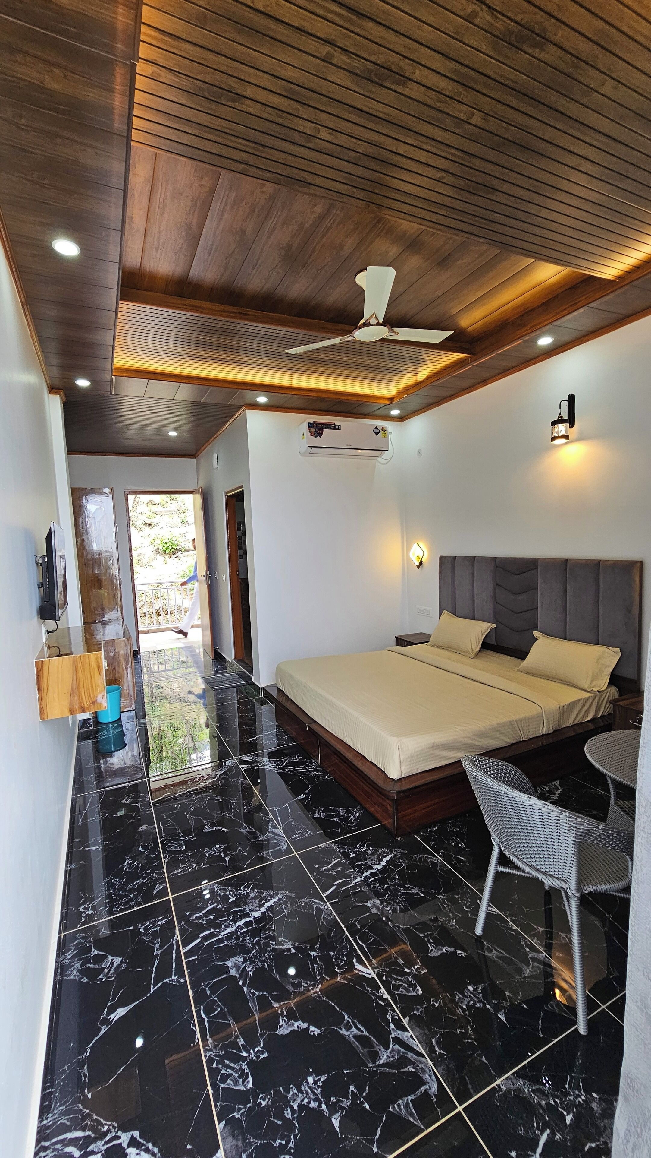 A bedroom with a bed, a ceiling fan, an air conditioning unit, a TV, and a bathroom with a toilet and sink.