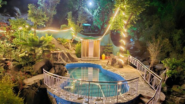 Une vue nocturne d’une piscine extérieure entourée de rochers et de végétation, avec un petit pont et un espace de repos abrité.