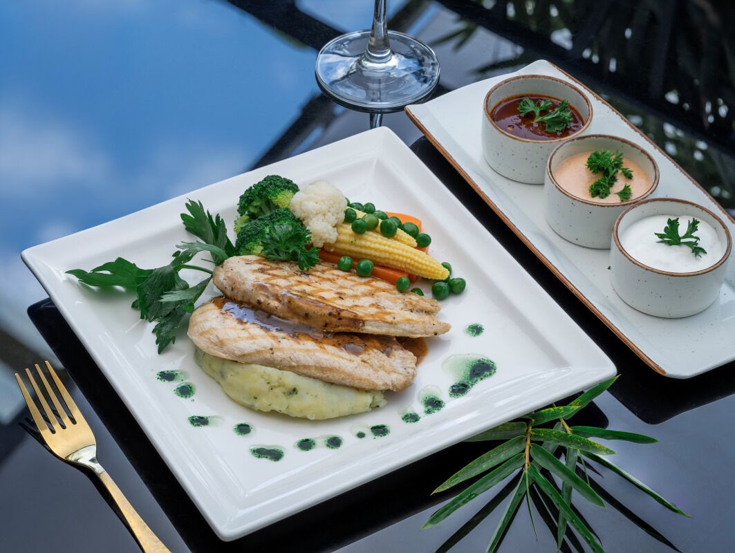 A white plate with grilled fish, vegetables, and mashed potatoes, accompanied by two small bowls of sauce.