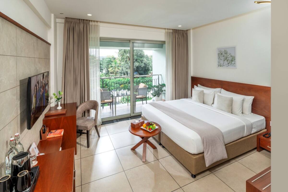 A hotel room with a large bed, a desk with a chair, a small table with fruit, and a balcony with outdoor furniture.