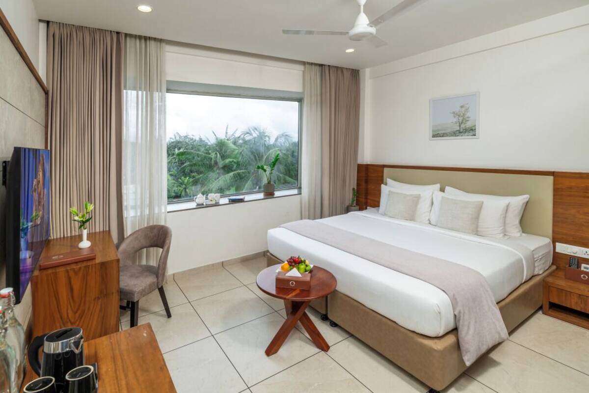 A hotel room with a large bed, a desk with a chair, a small table with a vase, and a window with curtains.