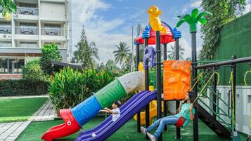 A playground with slides and swings, surrounded by greenery and a building in the background.